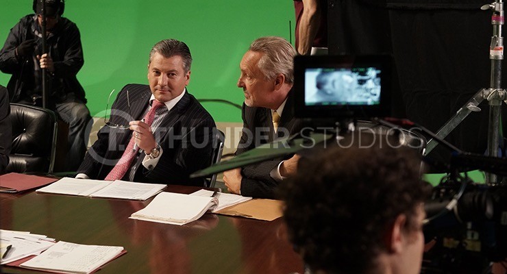 Inbetween shots during filming, Rodric David sits at at a boardroom table with a green screen backdrop.